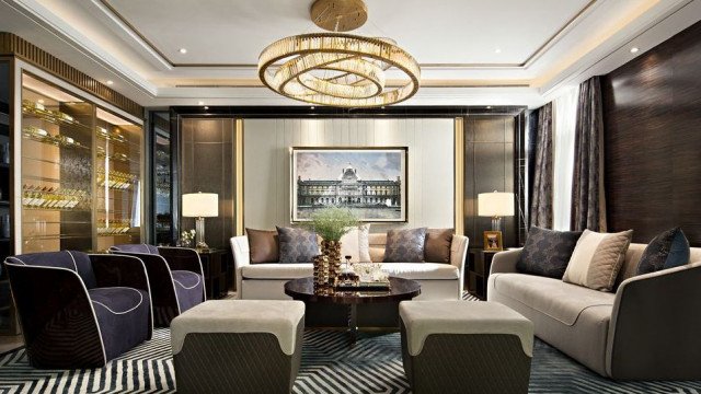 This picture shows a modern luxury style bedroom, with a unique and elegant interior design. The walls are painted in a light blue color, creating a calming atmosphere. On the left side of the room is a large bed with white bedding and decorative pillows. A wooden floor gives the space a warm and inviting feel. An ornately designed round night stand stands at the foot of the bed, and a comfortable armchair and ottoman sit by the window on the right side of the room. The area is illuminated by recessed lighting and a standing lamp near the bed. A beautiful