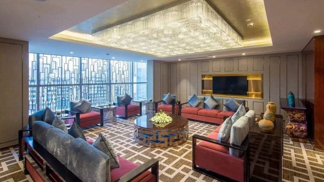 This picture shows a luxurious, contemporary living room with light gray walls and dark gray accents. The room features sleek white furniture with accent pillows, a large white framed mirror, and a few abstract art pieces. The center of the room is dominated by a grand crystal chandelier that hangs from the ceiling.