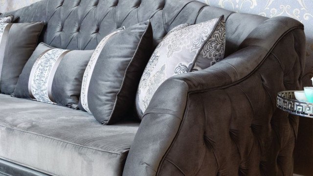 This picture is of a luxurious living room with a modern, minimalist design. The walls are painted a soft grey color and the furniture is white with gold accents. There is a large sectional sofa with a round ottoman in the center, two armchairs, an oversized coffee table, and several side tables. The rug in the center adds to the elegance of the room, and there is a large wall-mounted television on one wall. Large windows let in plenty of natural light and complete the look.