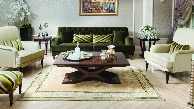 The picture shows a luxury living room with a beautiful modern interior design. The room features two luxurious sofas arranged in an "L" shape around a large center coffee table, and two armchairs with a footstool at the corner of the sofa. There is a large white leather ottoman in the middle of the room, surrounded by geometric patterned rugs. The walls are decorated with abstract art pieces and intricate moldings, while the ceiling is painted white with recessed lighting fixtures. The entire room is filled with modern and elegant furniture pieces, creating an inviting and luxurious