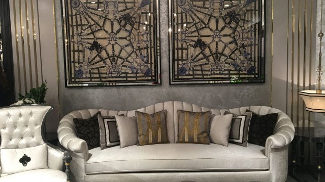 This picture shows a luxurious and modern living room. The room features a light gray wall with a white and black ornate pattern on one side. The center of the room is dominated by a large, white leather sofa and two navy velvet accent chairs. There is an elegant black glass chandelier hanging from the ceiling and several colorful throw pillows on the sofa. On one side of the room is an off-white, glass coffee table and a set of two burnt orange armchairs. On the other side of the room stands a tall, dark wood armoire for additional storage. The