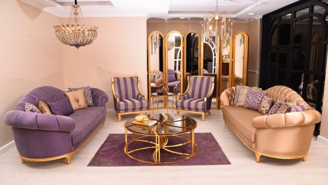 This picture shows a luxurious modern living room with a sleek and stylish design. The room is decorated with an ornate beige velvet couch and two plush armchairs with gold details. The walls are painted in a warm beige tone and feature an impressive abstract art piece. The room also has an intricate chandelier and a crystal coffee table, both giving the space an air of opulence.