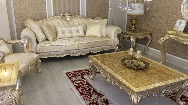 This picture shows a modern living room with luxurious furnishings. The walls and floors are covered in white marble and feature an ornate beige wall sconce. The primary seating is two beige sofas, flanking a white and gold coffee table. There are two armchairs with chevron-patterned backs, placed around the rug in the center of the room. On the opposing wall, there is a built-in shelf unit with glossy black cabinets and shelves. There is also a red chair with a glass side table beside it.