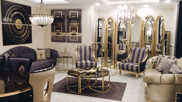 The picture shows an elegant contemporary living space with a large, curved gray velvet sofa against a backdrop of white walls and black accents. There is a rectangular glass coffee table in the center of the room. On the walls are abstract paintings in shades of red and purple for a pop of color. The room is filled with natural light from the tall windows behind the sofa.