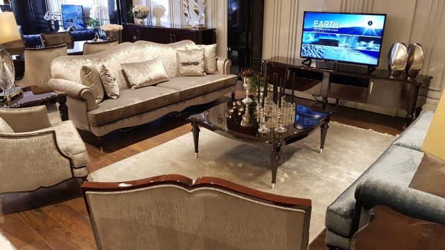This picture shows an elegant, luxurious living room with beige walls, large windows and light wood flooring. A white grand piano is situated in the center of the room, a modern art piece hanging on the wall behind it. An off-white sofa set with an ornate gold trim is positioned in front of the windows, a large matching ottoman in front of it and two ornate armchairs at the side. The room has several large, ornate mirrors and chandeliers, as well as a number of designer light fixtures.