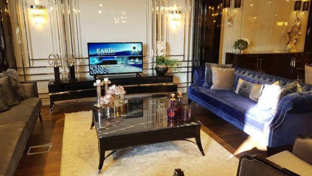 The picture shows an interior design project from Antonovich Design. The main feature of the room is a grand, curved staircase with ornate detailing. The staircase is surrounded by luxurious marble flooring and walls covered with a tapestry-like pattern. In the center of the room is an elegant crystal chandelier. The furniture surrounding the staircase includes plush couches, velvet armchairs, and an intricately carved coffee table.