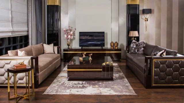 This picture shows a luxurious modern bedroom in beige and cream tones. It includes a large king-sized bed dressed with white and gold linens, a chandelier hanging from the high arched ceiling, two reading chairs in a corner, an ornate framed mirror on the wall, and an elegant rug in the middle of the floor.