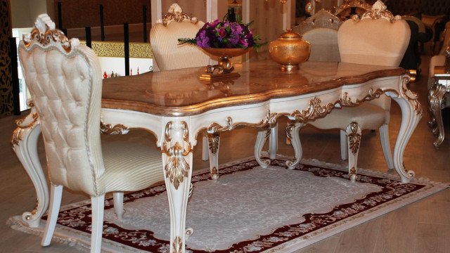 This picture shows a luxurious living room with a comfortable grey-colored sofa set, an ornate round table in the center and a large painting on the wall. The walls are painted a light mauve shade and the ceiling has intricate designs. There are also two arm chairs in the room and several pieces of decorative art on the shelves.