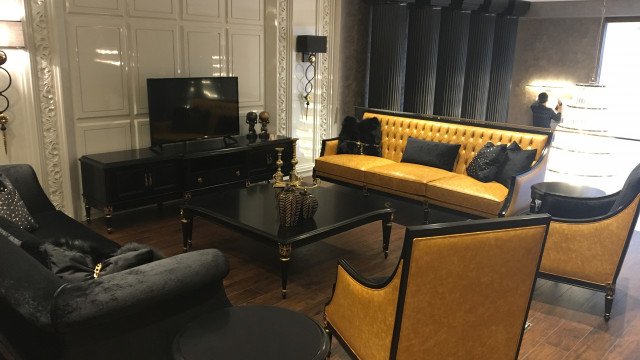 This picture shows a luxury living room. The room features a stunning gold chandelier suspended from the ceiling, a stylish fireplace with a large mantelpiece, and two tufted grey sofas with white and gold throw pillows. A leather steamer trunk serves as a coffee table, and there is an open bookshelf showcased in the corner. The walls are painted a soft white color and the floor is covered with a plush beige area rug.