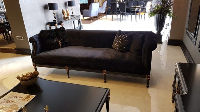 This picture shows a modern luxury living room designed by Antonovich Design. It features a white sofa lined with plush accent pillows, two white armless chairs, and an accent stool. The walls are painted a light grey and decorated with abstract artwork. A large rug is placed in the center of the room, and the floor is covered in dark-colored tiles. There is also a fireplace, a grand Chandelier, and a television mounted on the wall.