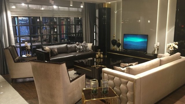 The picture shows a modern luxury living room designed by Antonovich Design, an interior design company in the United Arab Emirates. It features an elegant white leather couch with bronze decorative pillows, a matching white marble coffee table, and light hardwood floors. There is a sophisticated beige rug beneath the furniture, and a grand chandelier hanging from the ceiling to add a sense of drama and glamour to the space. The walls include wall art, framed photographs, a mirror, and window treatments.