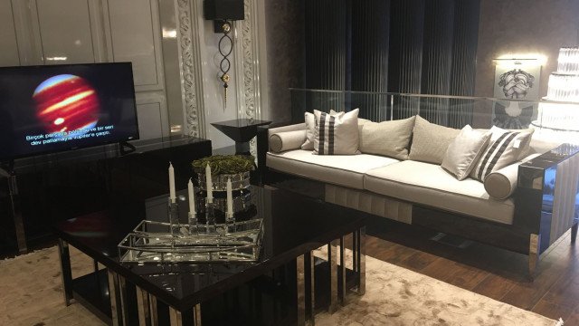 This picture shows a modern and luxurious living room with elegant brown and white furniture. The walls are painted in a light tan color and have a white crown molding framing the upper edge. There is a large framed mirror hanging on one wall, and a white sectional sofa with several large brown cushions arranged to form a U shape. The floor is made of dark wood, and a glass-topped coffee table is centered in the middle of the seating area. On the walls are several framed art pieces, and several potted plants give the room a cozy and inviting touch.