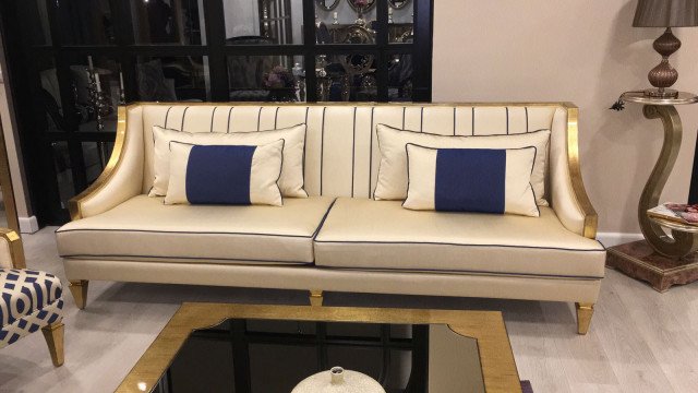 This picture shows an artfully designed living room, highlighting the use of color and texture. The room features a light blue velvet sofa with traditional patterned upholstery and silver accents. A pink armchair is placed in front of an abstract painting, and to the left is a grey tufted ottoman. The pale wood flooring is contrasted by a white shag rug and copper side table.