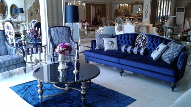 This picture shows a luxurious and modern living room. It features a built-in fireplace, off-white walls, and a deep navy-blue velvet sofa, with several accent pillows. The floor is light grey tile, and there are several area rugs in various shades of blue, as well as several plants for added warmth and texture. The large black framed mirror adds to the modern feel, while the colorful abstract painting on the wall provides a touch of artistry.