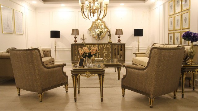 The picture shows an elegant living room that has a luxurious crystal chandelier hanging over a white and gold sofa. The walls are painted a light blue color, and the floor is covered with a white rug with intricate designs. There is also a small round glass and gold table in the center surrounded by white chairs. A few shelves with golden frames hang on the wall, displaying various decorative items.