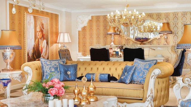 This picture shows an elegant and modern living room. The room features dark wood furnishings, a luxuriously tufted white leather sofa, a large round glass coffee table, two white armchairs, and off-white walls with stylish wall art. The room is decorated with several ornate lamps and vases. A large area rug helps to tie the entire look together.