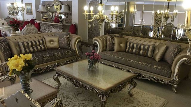 This picture shows a contemporary interior design of a large living room. The room features a beige suede L-shaped sectional sofa, a dark gray accent wall, and a striking crystal chandelier hung from the ceiling to create a luxurious and modern look. The room also includes an ornate area rug, a collection of gold and white accent pieces, a fireplace, and several large windows that provide plenty of natural lighting.