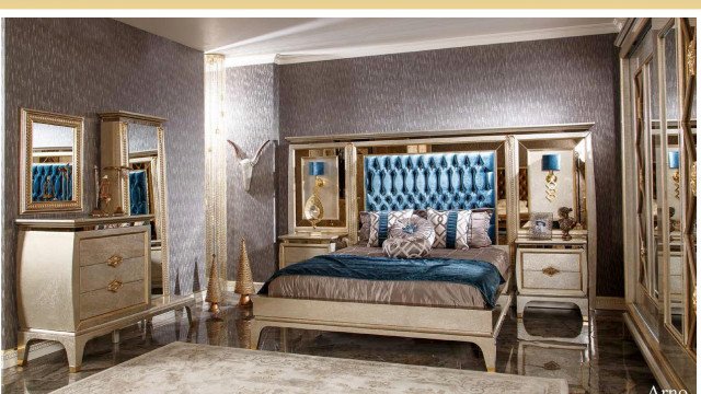 This picture shows a luxurious bedroom designed by Antonovich Design. The room has an ornate design with a white and cream colour palette. The walls are adorned with a sophisticated wallpaper, while the floor is covered with a white marble tiled design. There is an elegant four-poster bed at the centre of the room, surrounded by two large chairs and a luxurious chaise longue. A grand crystal chandelier hangs from the ceiling, while a fireplace is built into one of the walls. An intricate rug lies in the centre of the room, adding a touch of warmth to the