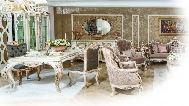 This picture shows the interior of a luxurious living room. The walls are covered in a pale pink fabric, and the floor is tiled with a marble pattern. The walls are adorned with golden detailing, and there are two large windows that filter light through sheer fabric curtains. An antique-style sofa sits in the center of the room, and a glass coffee table holds a few decorative accents. A crystal chandelier hangs from the fan-shaped ceiling and casts a gentle light throughout the room.