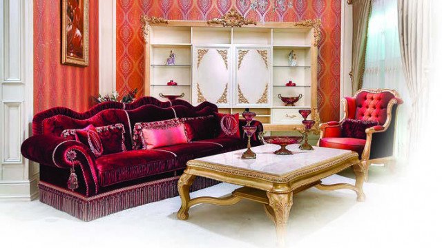 This picture is of an elegantly designed living room in a luxurious home. It features a white leather sofa and armchair with a white ottoman, a large grey area rug, an ornate floor lamp with a white shade, and a glass coffee table. The walls are painted in a light grey color, while the ceiling boasts an intricate white design. Large windows let in plenty of natural light, while a large candle-style chandelier hangs from the center of the ceiling.