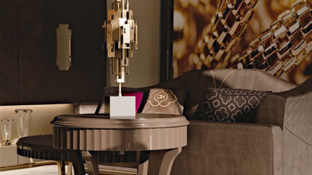 This picture shows a contemporary luxury living room and dining area design by Antonovich Design. The room features a beige and white color palette with accents of gold, which is seen in the light fixtures, furniture, wallpaper, and decor throughout. A large cream sofa and two armchairs create a cozy seating area around the stone fireplace. The dining area is separated by semi-transparent curtains and boasts a beautiful glass table and chairs. Two round gold chandeliers hang over the table, completing the look of this sophisticated and stylish space.