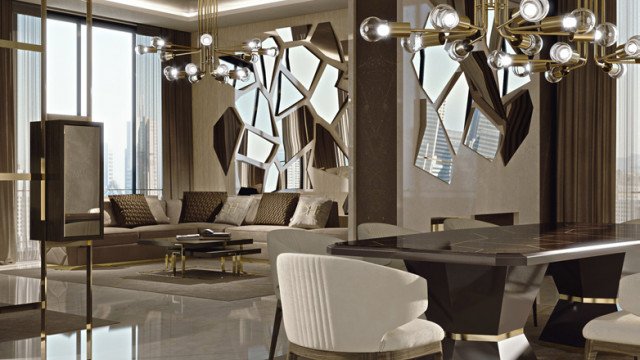 This image is of a modern interior living room designed by Antonovich Design. It features a sleek grey sofa with two white armchairs, a glass coffee table, and a textured grey rug. The room has multiple sources of lighting, including up-lighting around the perimeter of the room, a modern hanging chandelier, and additional floor lamps. Grey wall panels are adorned with framed artwork to add texture and interest to the space.