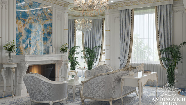 This picture shows a beautiful modern bedroom with high-end luxury furniture and decor. The centerpiece of the room is the large bed which features intricate tufted headboard and bed frame upholstered in a light beige fabric. On either side of the bed are two nightstands with contemporary glass lamps on top. In the corner is a sleek white armchair paired with a modern ottoman with gold accents. There is also a statement mirror on the wall with an ornate wooden frame. Above the bed is a large abstract art piece adding a bit of colour to the room