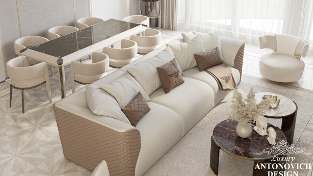 This picture shows a contemporary interior design created by Antonovich Design. It features a pale grey sectional sofa along with two armchairs arranged to form an "L" shape. The entire furniture set is placed on a plush, light-toned area rug that provides a unified look and creates a cozy atmosphere in the room. The seating area is also surrounded by a white wall with wood finishes and several artwork pieces. On the side, there is a modern glass coffee table with a sleek metal frame, adding a touch of sophistication to the space.