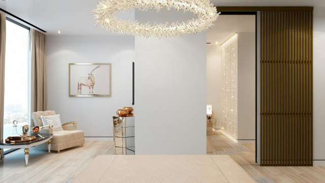 This picture shows a modern living room with luxurious accents. The space is decorated with a light gray velvet couch, a glass coffee table, a colorful area rug, and an ottoman. There is also a stylish accent wall featuring cascading greenery, giving the room a unique touch. The walls are painted a neutral color, and the hardwood floors bring warmth to the space. Polished chrome fixtures and gold accents add a touch of glamour, while contemporary artwork gives the space a modern edge.