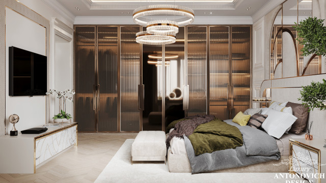 This picture shows a luxurious bedroom designed by Antonovich Design. The room has a modern and elegant aesthetic, featuring marble walls and floors in muted tones paired with warm wood accents. A large window allows natural light to flood into the space and the light furniture pieces give the room an airy feel. In the center of the room is a stunning four-poster bed draped in blue and white fabric and decorated with tassels and pillows. On either side of the bed are two ornate nightstands adorned with golden fixtures, while a chaise lounge adds additional seating to complete the
