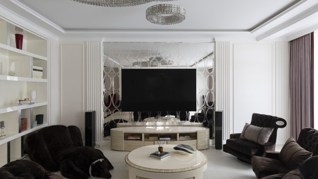 This picture is of a luxurious and modern living room. The room is decorated in various shades of white and beige and features a few accent pieces that add a touch of color. A large sectional couch sits in the center of the room, allowing for plenty of room to relax and entertain guests. In the corner of the room there is a plush cream-colored area rug that creates a cozy spot for conversation and lounging. The walls are adorned with several framed artwork pieces, giving the room an elegant touch. There is also a glass coffee table and a sleek black chandelier