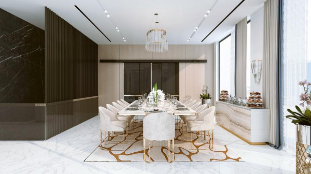 This picture shows a luxurious, modern kitchen with two islands. The countertops are white marble, and the cabinets are a light, wood grain with brushed gold accents. The appliances are stainless steel, and the backsplash is textured tile. There is recessed lighting and a large window, as well as additional pendant lights above the islands. There is seating area near one of the islands.