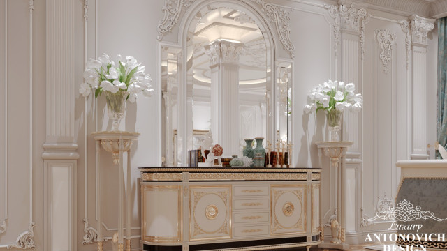 This picture shows a luxurious, grand entrance to what appears to be a hotel or residence. The entryway is made up of an ornate wood and glass door, flanked by white stone pillars adorned with intricate carvings. The walls are draped with thick gold curtains, and the hallway is carpeted in a rich, deep royal-blue color. There is a double staircase leading up to the door, lined with a glowing golden railing. A crystal chandelier hangs from the ceiling, creating a warm and inviting atmosphere.