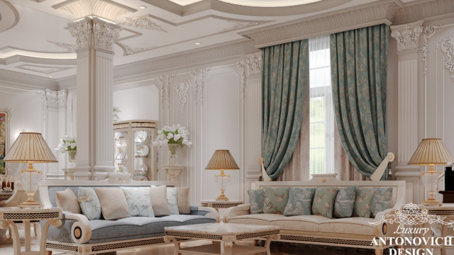 This picture shows a modern bedroom designed by Antonovich Design. The room has a minimalist style, with simple white walls and a light hardwood floor. The bed is covered with a light beige blanket and pillows and backed against a headboard which has an intricate floral pattern. There is a window to the side which is framed with cream drapes, and a white armchair sits in the corner of the room. On the wall opposite the bed is a large painting of lush green foliage.