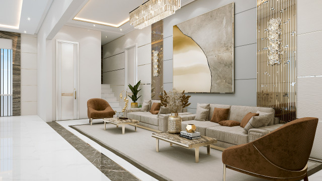 This picture shows a large and luxurious living area. The room has cream-colored walls and ceiling, with golden trimmings on the ceiling, cornices, and doorways. In the center of the room is an elegant grey sofa with two white armchairs and a round coffee table. On the left side of the room are two large windows letting in natural light and framing a view of trees and greenery outside. On the right side is a tall wooden cabinet with several shelves, as well as a flat-screen television on top. The floor is a light brown wood laminate with