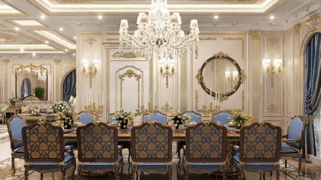 The picture shows a luxurious bedroom in an ornate style. The room features a large canopy bed with a velvet headboard, footboard and draperies in shades of gold and burgundy. Pristine white walls are framed by intricately carved moldings, and the ceiling displays beautiful stucco details. A golden chandelier and two elegant side tables adorn the room, along with plush velvet armchairs in the corner. In the background, a large window is covered with dramatic drapery that extends to the floor.