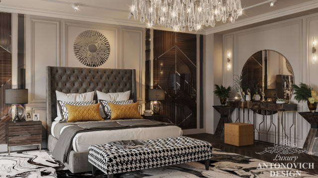 This picture shows a luxurious master bedroom decorated in an elegant modern style. The room features a beautiful four-post bed with a headboard and footboard encrusted with crystals, a velvet tufted ottoman at the foot of the bed, and an ornately designed chandelier hanging from the ceiling. Wall sconces flank each side of the bed and an area rug anchors the space. The walls are painted a soft grey, while the trim is white, giving the room an airy feel.