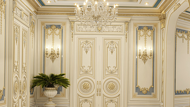 This picture shows a luxurious grand salon in a modern, upscale home. The room features a large white and gold sofa with two velvet ottomans on either side, as well as two matching white and gold armchairs. A heavily ornate gold and crystal chandelier hangs from the center of the room, and a white-and-gold carpet covers the floor. An intricate wall design of gold, cream, and white accents complete the look of the grand salon.