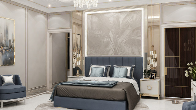 This picture shows a luxurious bedroom designed with modern decor. There is a large bed with a white and gold headboard, two beige armchair with gold accent cushions, a mirrored vanity table with a marble top, and a lounger with an ottoman on the side. The whole room is decorated with light neutral hues and intricate wallpaper designs, creating an inviting and cozy atmosphere.