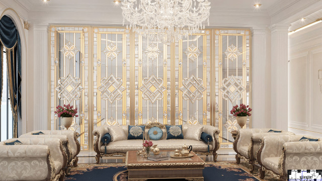 This picture shows the interior of a modern and luxurious living room with beige and cream-colored walls, hardwood floors, and a large, ornate white sofa. The furniture and accessories in the room create a warm and inviting atmosphere, with a gold accents throughout. On the left side of the room is an artistic accent wall featuring a set of large, elegant paintings, as well as several pieces of art hung on the wall. On the right side of the room is a fireplace, which adds to the cozy ambiance, and a television mounted above it for entertainment.