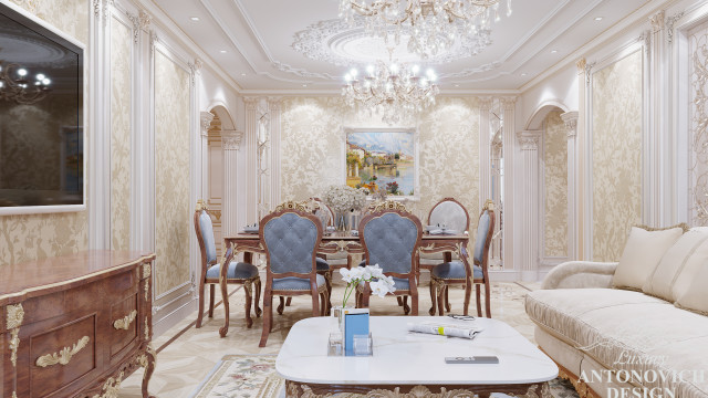 This picture shows a luxurious living room designed for entertaining and relaxation. The space features a large beige-colored sofa set up in the center, with two matching light brown armchairs and an accent chair off to one side. A glossy black coffee table sits in front of the furniture, and a modern patterned rug adds a decorative touch to the room. A glass chandelier hangs overhead and wall art pieces add to the luxurious feel of the space.