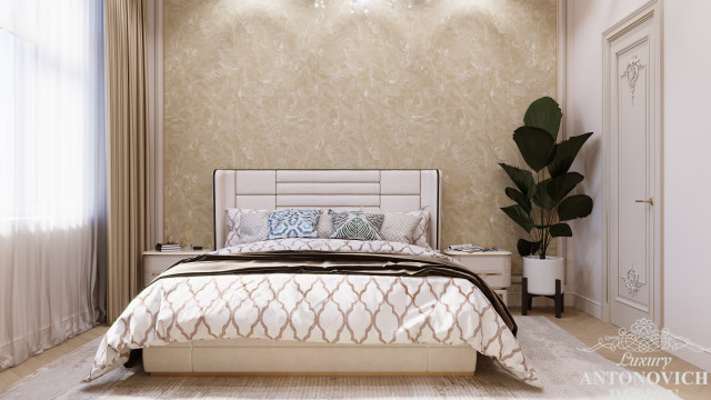 This picture shows a luxury bedroom decorated with an elegant, modern design. The room features a large bed with a white headboard, patterned white and gray bedding, and many matching accent pillows. The room is accented with a marble-topped night stand and a vibrant pink accent chair with gold upholstery. A metal wall sconce and candles with gold holders also add to the decor. The walls are painted a coordinating shade of light gray and have several pieces of art hung on them. Lastly, a plush white area rug completes the look.