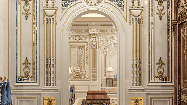 This picture is of a large, luxurious interior space. It features a grand, curved staircase with gold and marble accents and a luxurious chandelier hanging from the ceiling. There are also two tan chairs placed in front of the staircase, flanked by two white columns. The walls are painted a light color and decorated with ornamental, golden molding. There is also a large mirror, with an intricate golden frame, situated between the two chairs.