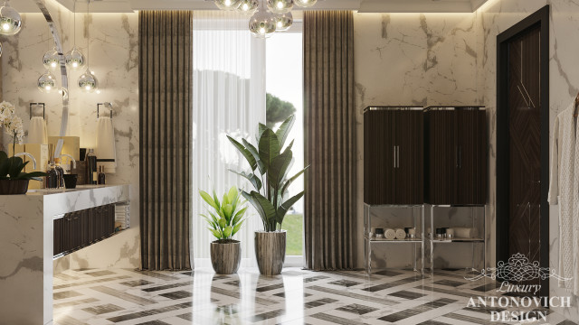 This picture shows a luxurious bedroom interior design. The walls and ceiling feature intricate, ivory-colored wall paneling and a high, coffered ceiling with stylish designs. The room contains two ornate night stands and a matching four-poster bed with an upholstered headboard, both in an elegant gold finish. A large, framed wall mirror adds a sense of light and space to the room, while two regal armchairs upholstered in a bold floral fabric complete the luxurious look.