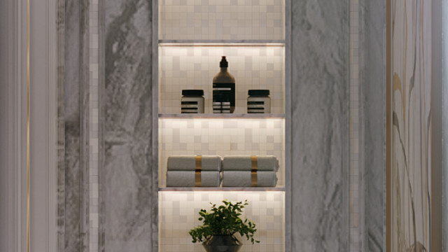 This picture shows a modern home office space in a rich and luxurious design. The walls are painted in a light gray hue and feature intricate gold-colored wall sconces. The desk is placed in the middle of the room and is made of glass and black metal accents. On either side of the desk, two large windows let in plenty of sunlight, as well as two blue armchairs for additional seating. The room is decorated with framed art and various plants, creating a relaxing atmosphere.