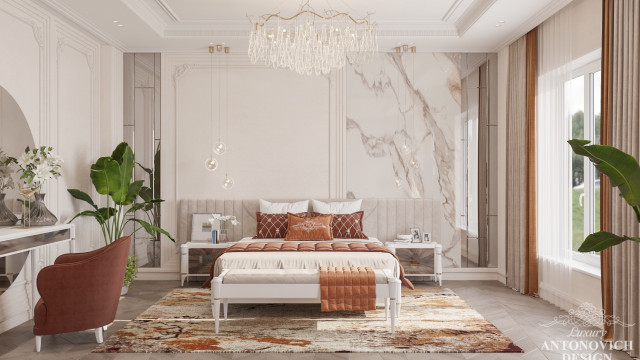 This picture shows a luxurious bedroom with modern design elements. The walls are painted a neutral light gray to emphasize the white and silver furniture. The headboard is made of a curved white leather material and is accompanied by white nightstands. The bed is topped with several silver throw pillows in various sizes, as well as a blanket that has a decorative metallic pattern on it. Mirrored accents and a tall lamp on either side of the bed complete the look.