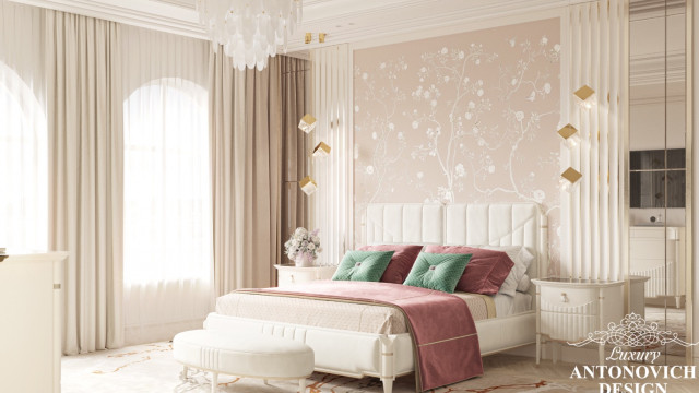 This picture is of a luxurious bedroom designed by Antonovich Design. It features a tufted headboard and bed frame in off white, complemented by cream-colored walls and flooring. The room also has a large crystal chandelier, ornately carved wooden furniture pieces, a window with rich drapes, and multiple soft pillows on the bed. The overall effect is one of opulence and sophistication.
