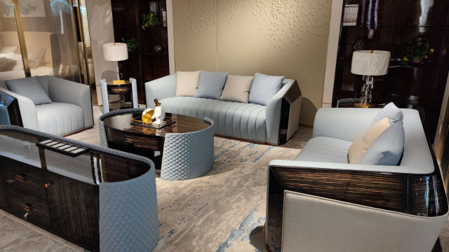 This picture shows a modern, luxurious living room with grey-black decor. The walls are painted a dark colour, and the floor has a light-grey rug beneath a white velvet couch with bright yellow pillows. There is a white coffee table in the centre of the room, and on either side of the table are two black leather armchairs. On the wall behind the couch, there is an abstract painting in shades of black and white. Above the couch, a large golden sunburst mirror hangs on the wall. The room is lit by several statement lighting fixtures which add to the modern