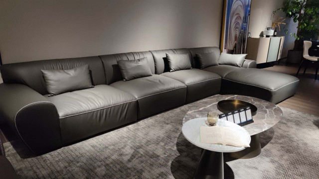 This picture shows a modern luxury bedroom design. The walls are painted a neutral beige color, and the floors are covered in a soft cream-colored rug. The ceiling is adorned with recessed lighting, and the bed has an upholstered headboard. To the left of the bed is a tall dresser, and to the right is a cozy lounge chair. At the foot of the bed is a white accent table with a vase of fresh flowers. On the wall behind the bed is a large abstract painting.