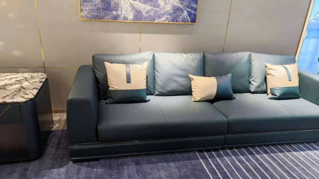 This picture shows a living room with modern luxury furniture design. A luxurious blue velvet sofa is placed in the center of the room and it is surrounded by a white coffee table, two accent chairs, and a decorative rug. The walls are a light gray color with a subtle geometric pattern and a few large abstract paintings for decoration. On one wall there is a shelving unit that holds a selection of books, photo frames, and other decor items. The flooring consists of a light wood laminate and there are several large windows that allow plenty of natural sunlight into the space.